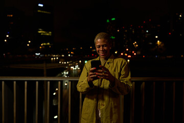 Woman smiling using phone on urban night balcony