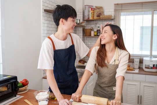 Asian couple wearing aprons and smiling, baking bread in their kitchen. Man showing affection to woman while cooking
