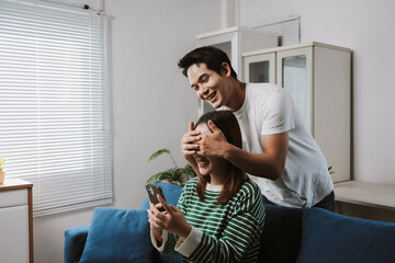 Young man playfully surprising his girlfriend by covering her eyes while she is using a smartphone at home