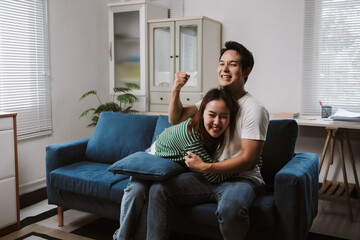 Obraz premium Happy Asian couple on a sofa in a modern living room, cheering and celebrating together while watching TV and enjoying weekend relaxation