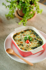 Delicious spinach bake with melted cheese, served in a red baking dish on a plate with a wooden spoon, garnished with a tomato and a plant background