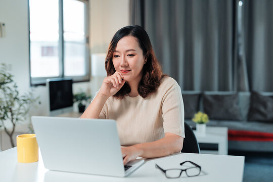 Asian woman concentrating on work while typing on laptop keyboard at home office desk, embracing freelance productivity