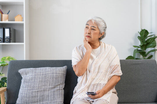 Senior Asian woman sitting on a couch, experiencing stress, loneliness, and confusion while holding a smartphone - Powered by Adobe