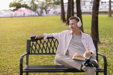Obraz premium Mature man enjoying leisure time on a park bench, listening to music with headphones, holding a coffee cup, and reading a book on a sunny day outdoors