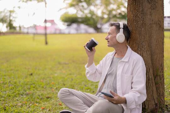 Mature man sitting on grass in a park, leaning against a tree, sipping coffee from a disposable cup, and listening to music through headphones while holding a smartphone
