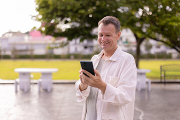 Mature man standing in a city park, smiling as he uses his smartphone for messaging and browsing, relaxed and enjoying digital connection in a sunny outdoor setting