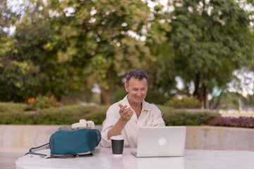 Senior man smiling during a laptop video call while working remotely at a park table with coffee and bag, enjoying flexible freelance life and outdoor connectivity