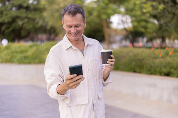 Senior man standing outdoors in a park, happily engaging with his smartphone, enjoying a hot coffee, representing modern technology use and relaxed leisure for older adults