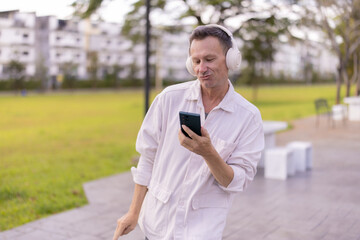 Mature man relaxing in a sunny urban park, smiling while streaming music on wireless headphones and browsing content on his smartphone, enjoying digital leisure outdoors