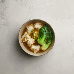 Wonton soup with bok choy in dark bowl on light background, top view