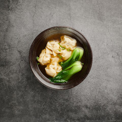 Wonton soup with bok choy in dark bowl on light background, top view