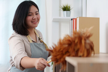 Home Cleaning. A woman dusting shelves with a feather duster, showcasing a tidy living space.