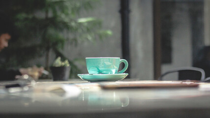 Bright turquoise tea cup with matching saucer sits on a wooden table in a calm outdoor cafe. Soft...