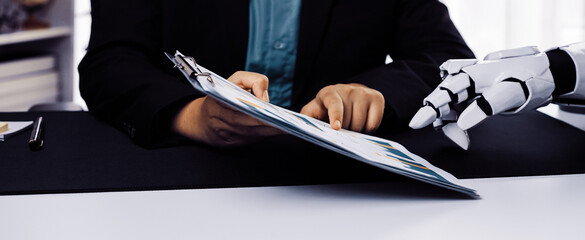 A human hand reviews financial documents with the assistance of a robotic hand in a modern office setting, highlighting technological advancements in the corporate world.Noogenesis