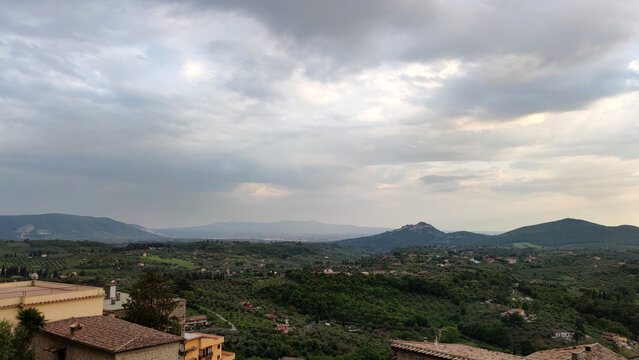 A wide landscape view of the Italian countryside with rolling green hills, olive groves, and small village houses under a dramatic cloudy sky