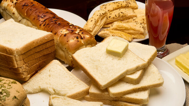 A close-up image of assorted freshly baked breads arranged on a breakfast table. The assortment includes sliced white bread, garlic loaf, puff sticks, and a cube of butter on a slice - Powered by Adobe