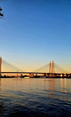The scene captures a large, modern bridge spanning the Neva River; nobody is directly visible. The setting sun casts a strong, beautiful golden light across the water and the distant city.