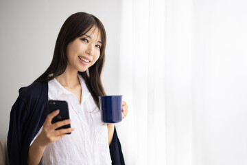 Young Woman Using Smartphone While Holding a Mug by the Window