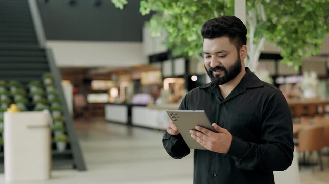 Portrait of happy Indian man scrolls internet on digital tablet in office building. Smiling bearded entrepreneur checks social media feed on mobile device in corporate coworking - Powered by Adobe