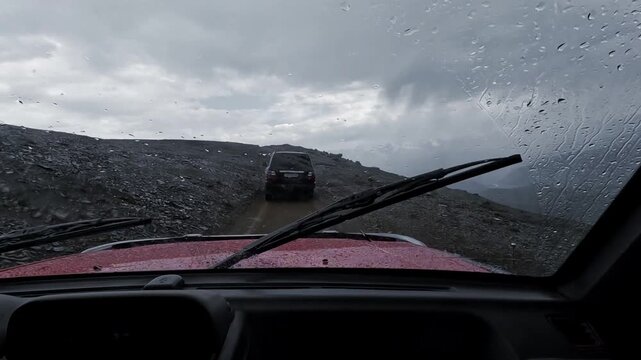 Mountain primer in the rain: view from the interior of an SUV through the windshield with drops and wipers; car in front, the feeling of an expedition.
