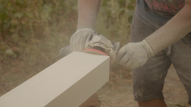 A worker is thoroughly sanding wooden beams using a polished grinder in an outdoor environment. Dust flies as they focus on creating a smooth surface.