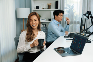 Profile smiling beautiful Asian businesswoman looking camera while holding cup to pose at modern office on working desk. Blurry background man colleague analyzing data market report on pc. Infobahn.