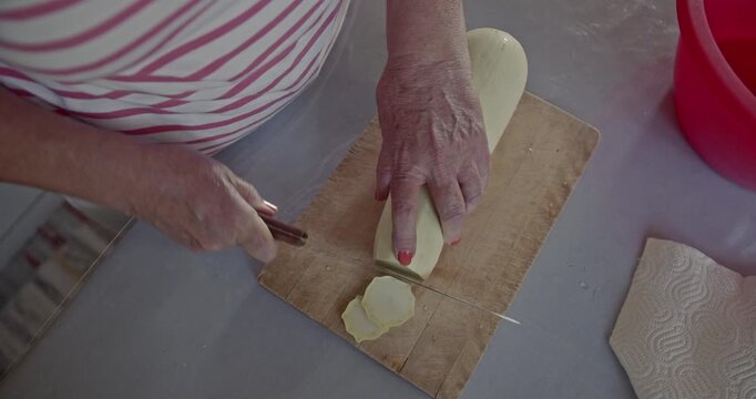 Close-up of an elderly woman's hands carefully slicing fresh zucchini into thin rounds on a rustic wooden cutting board. A red bowl waits for the healthy vegetable pieces.