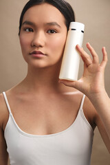 asian woman holds a white bottle in a simple studio setting, focused expression, beauty product...