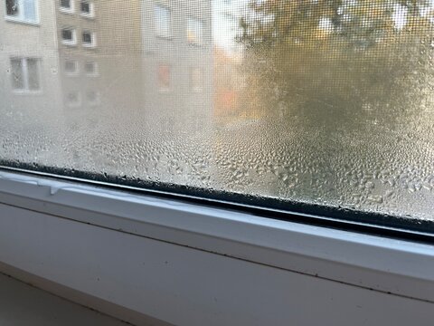 Close-up photo of condensation and water droplets on window glass with blurred apartment in autumn landscape in the background. Great for weather, home comfort, or seasonal mood imagery