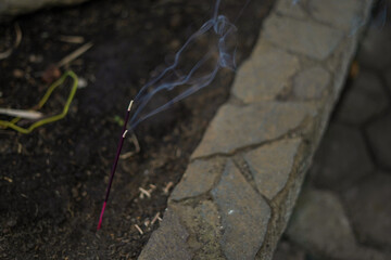 A single stick of burning incense smolders in the ground, releasing delicate wisps of smoke next to a stone border.