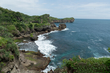 A scenic view of a rugged coastline with green cliffs meeting the deep blue ocean and white waves crashing against the rocky shore under a bright sky.