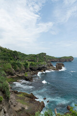 Coastal landscape featuring rocky cliffs, lush green vegetation, and the vast, blue ocean under a partly cloudy sky.