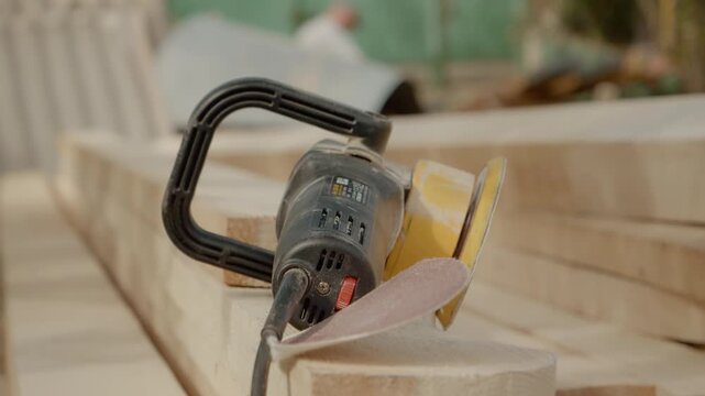 A random orbital sander sits on wooden beams, covered in dust, after completing the sanding process. The area shows evidence of recent woodworking activity.