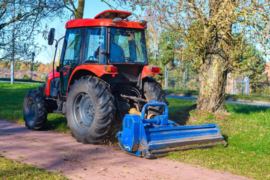 Red tractor mowing grass with blue flail mower near tree in park
Red agricultural tractor with a blue flail mower attachment cutting grass near a tree on a sunny day. 