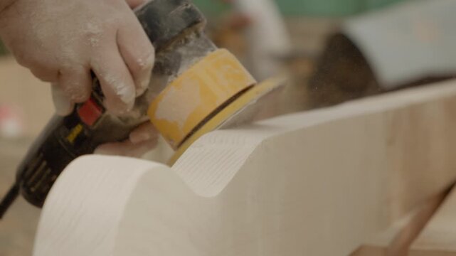 A worker carefully sands wooden beams using a random orbital sander. Dust fills the air as the tool smooths rough edges on the wood in a bright workspace.