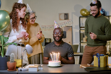 Middle aged Black man sitting at desk smiling with birthday cake and candles while young Caucasian woman, senior Caucasian woman, and young Caucasian man standing nearby wearing party hats