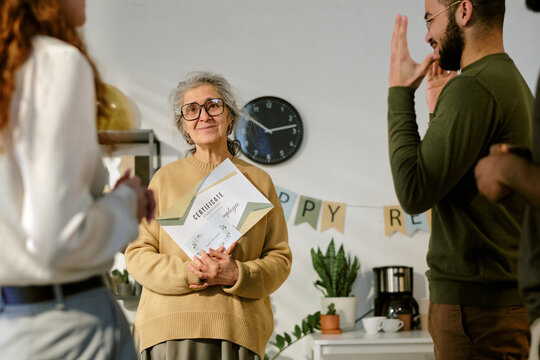 Portrait of senior Caucasian woman holding certificate while standing among diverse group of young adults congratulating her during celebration in modern indoor setting