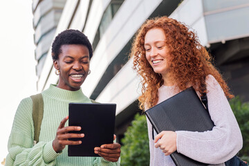 two young women smiling while using a tablet together in front of a modern building, one of them holding a folder, concept of friendship and student lifestyle