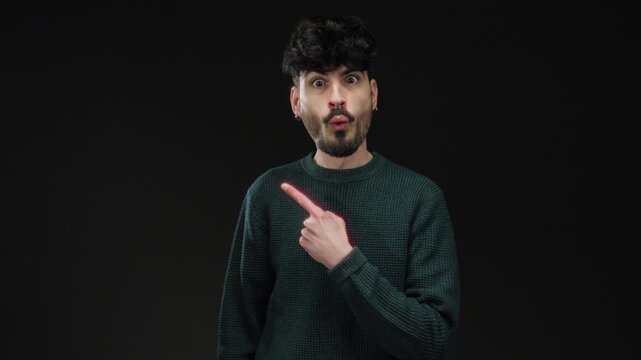 Young man in green sweater with surprised expression points to side against dark background conveying curiosity and intrigue.