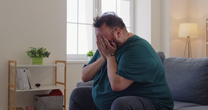 Tired fat overweight obese man holds forehead on couch. Male person with obesity shows headache, fatigue, stress, pain, migraine, exhaustion, and worry while sitting on sofa at home