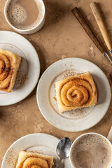 Freshly baked cinnabon roll buns served on ceramic plates with coffee on brown textured background. Cozy autumn breakfast concept. Top view