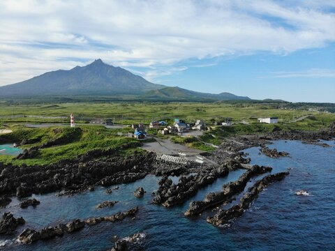 Coastal village and lighthouse beneath Mount Rishiri, rugged basalt reefs and blue Sea of Japan, Rishiri Island, Hokkaido, Japan — aerial drone view - Powered by Adobe