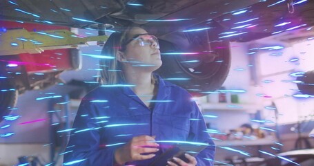 Asian mechanic wearing coverall inspecting undercarriage under lift in workshop, with tablet