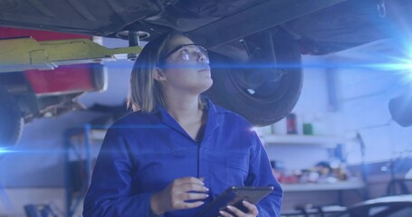 Female automotive technician examining undercarriage in repair workshop, with tablet, tool shelves