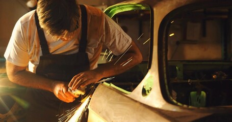 Auto body technician in overalls grinding stripped car frame in workshop, with angle grinder sparks