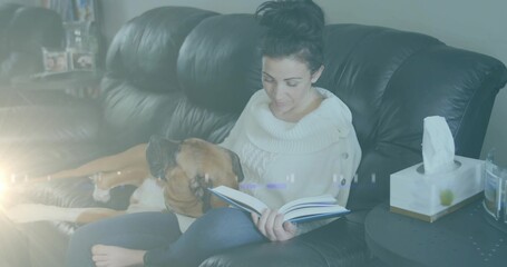 Reading woman in white sweater petting dog on black couch in living room, with open book