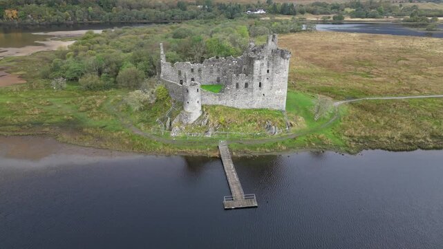 Aerial view of the Kilchurn Castle's ruins with a wooden pier extending into the loch, contrasting with green grass, Scotland.