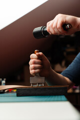 Crafting belt holes with punch tool on leather strap. Punching and detailing leather belt holes. Artisan punching holes into a leather strap for a handmade belt on a green cutting mat.