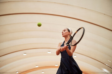 Young adult Caucasian woman swinging tennis racket indoors, focusing on hitting tennis ball during practice session, athletic movement under curved ceiling structure