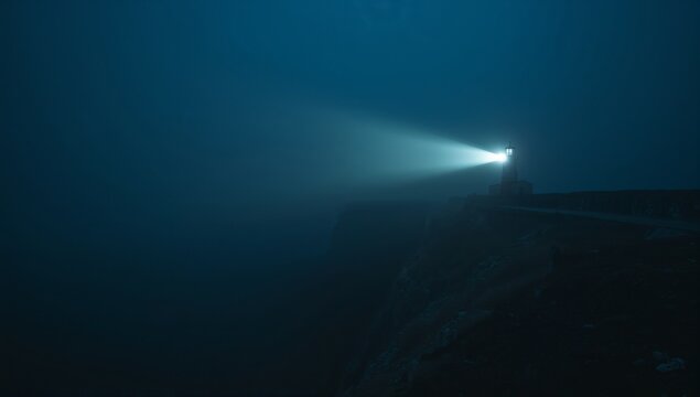 Shining lighthouse sending beacon through fog on rocky cliff at night, with narrow path, copy space